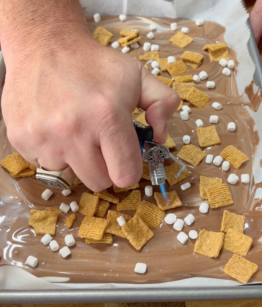 A hand uses a small kitchen torch to toast mini marshmallows on a tray of melted chocolate and cereal pieces.