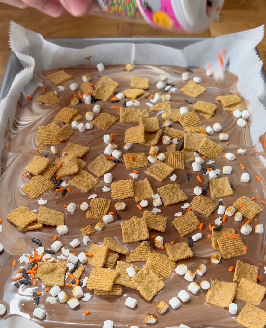 A baking sheet lined with parchment holds melted chocolate swirled with white chocolate, topped with cereal squares, mini marshmallows, and orange, black, and white sprinkles.