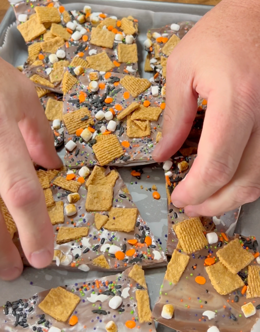 Hands break apart pieces of chocolate bark topped with cereal squares, mini marshmallows, orange sprinkles, and colorful edible confetti on a parchment-lined baking sheet.