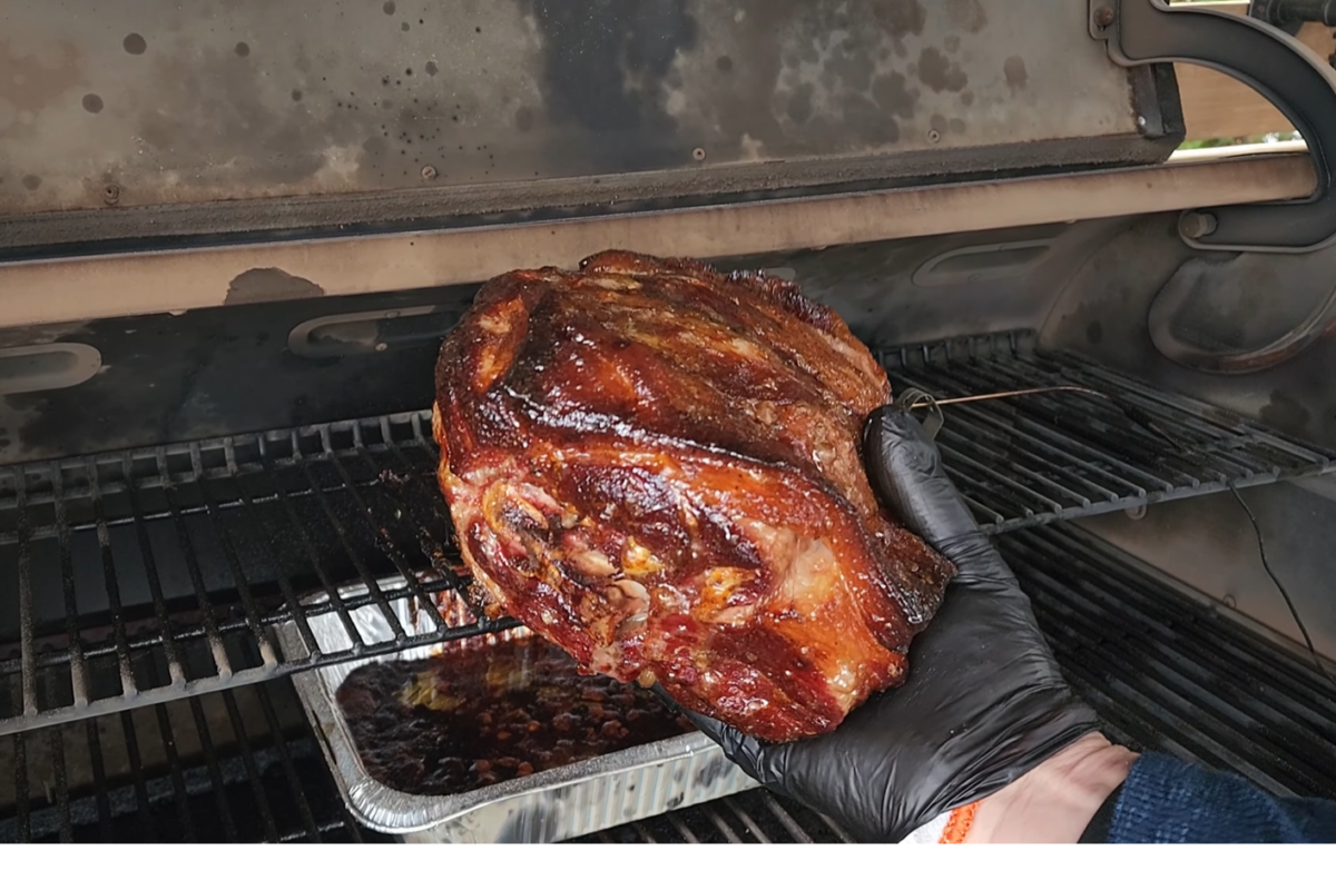 A gloved hand holds a large, cooked piece of meat inside a smoker grill, with a drip pan underneath on a lower rack.