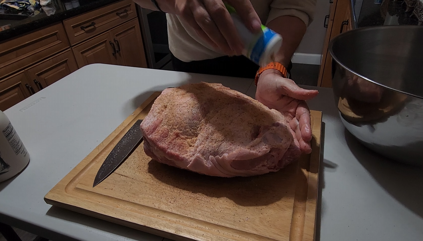 A person seasons a large piece of raw meat on a wooden cutting board, with a knife nearby and a metal bowl on the table.