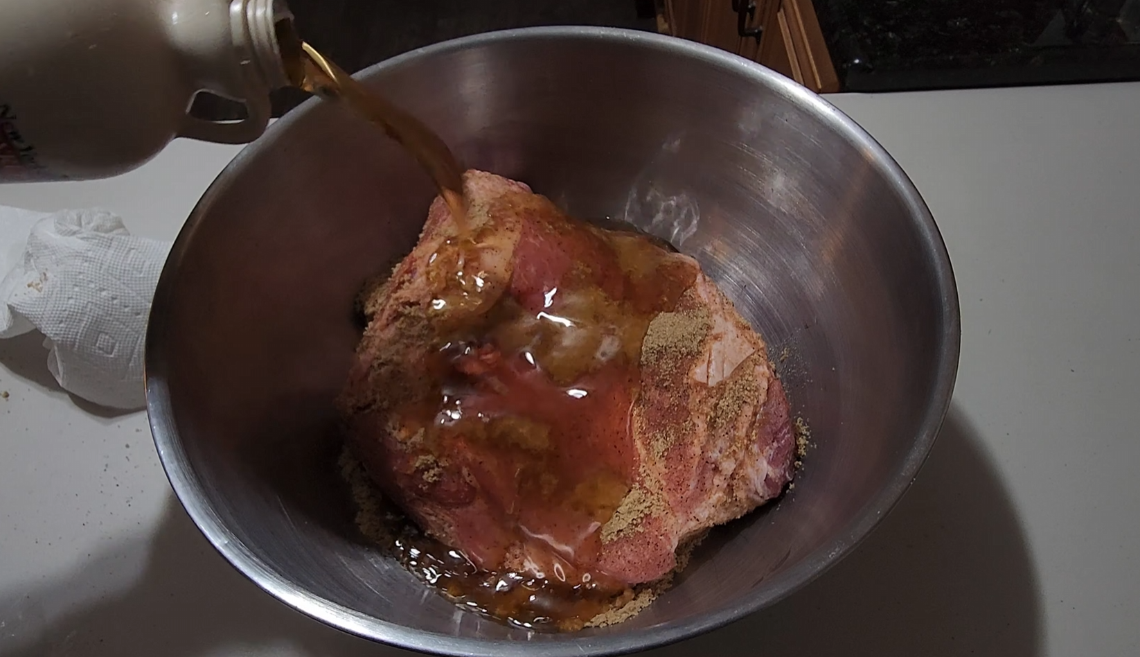 A brown liquid is being poured from a bottle onto a seasoned raw pork roast in a large metal mixing bowl on a kitchen counter.