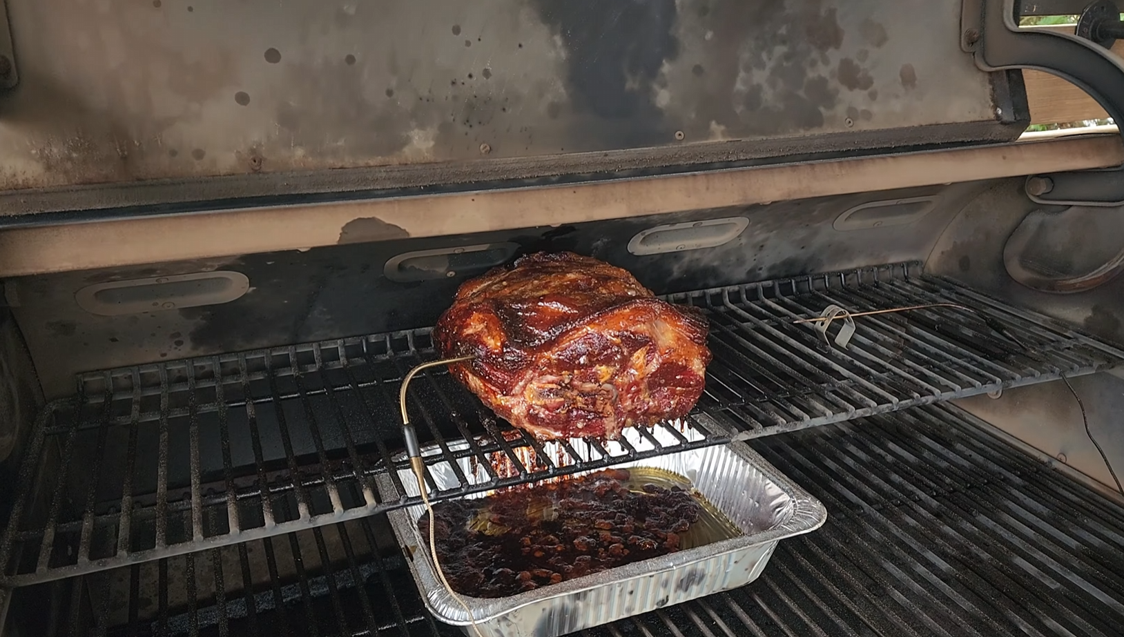 A large piece of seasoned meat is roasting on a grill rack, with a thermometer probe inserted. A foil tray below catches drippings and juices. The inside of the grill is dark and slightly stained.