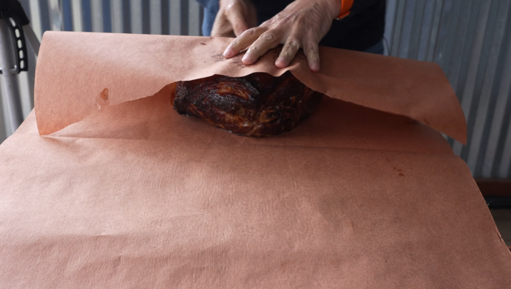 Person wrapping a smoked meat brisket in large brown butcher paper on a table, with hands folding the paper over the meat.