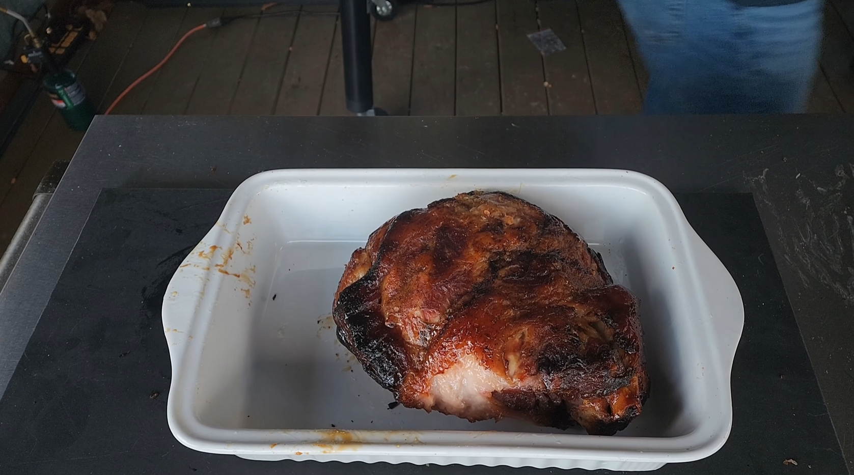 A cooked, browned roast sits in a white rectangular baking dish on a dark countertop; part of a wooden deck and a persons leg are visible in the background.