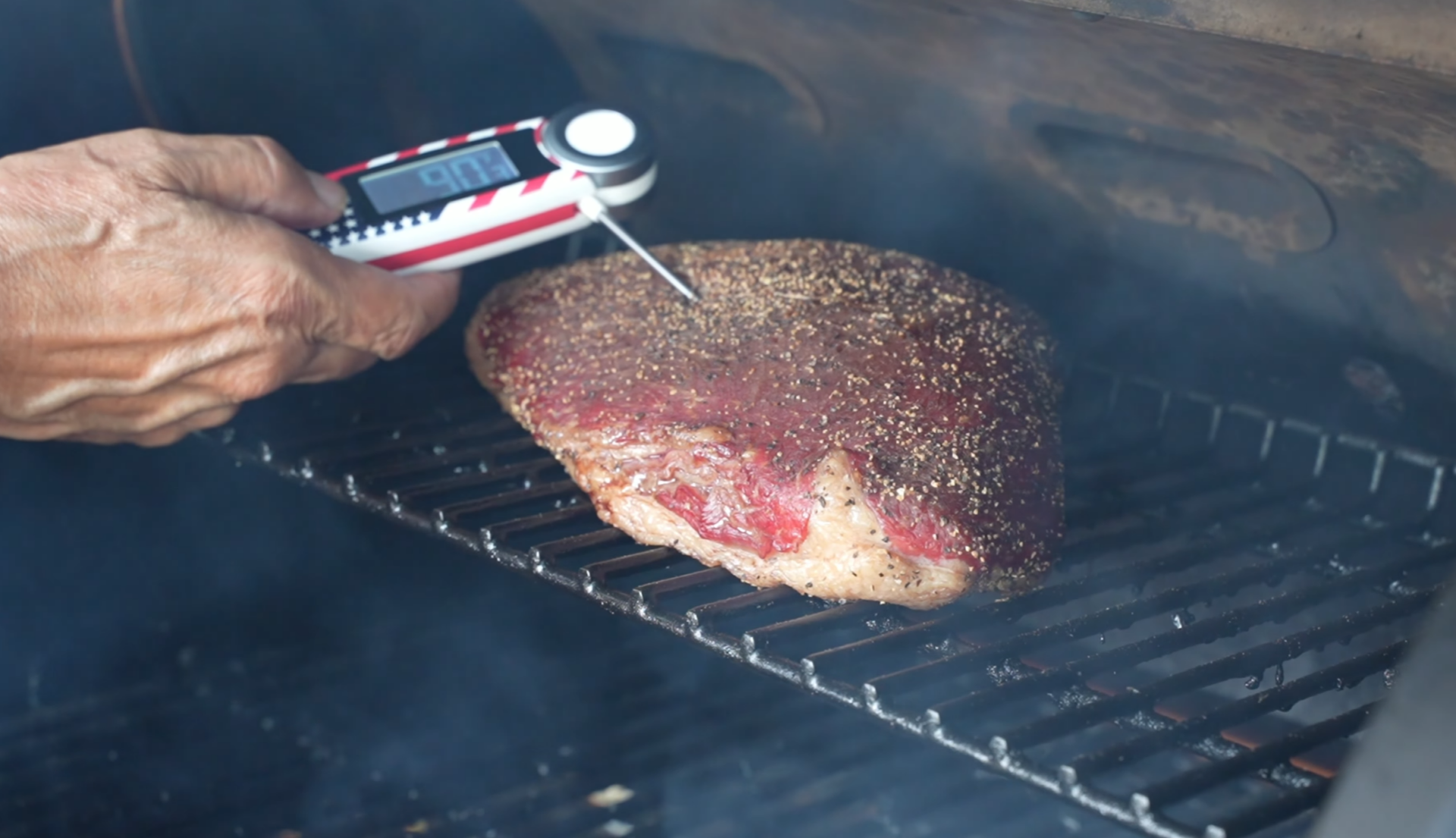 A hand checks the temperature of a seasoned beef brisket using a digital meat thermometer while it cooks on a grill with visible smoke.