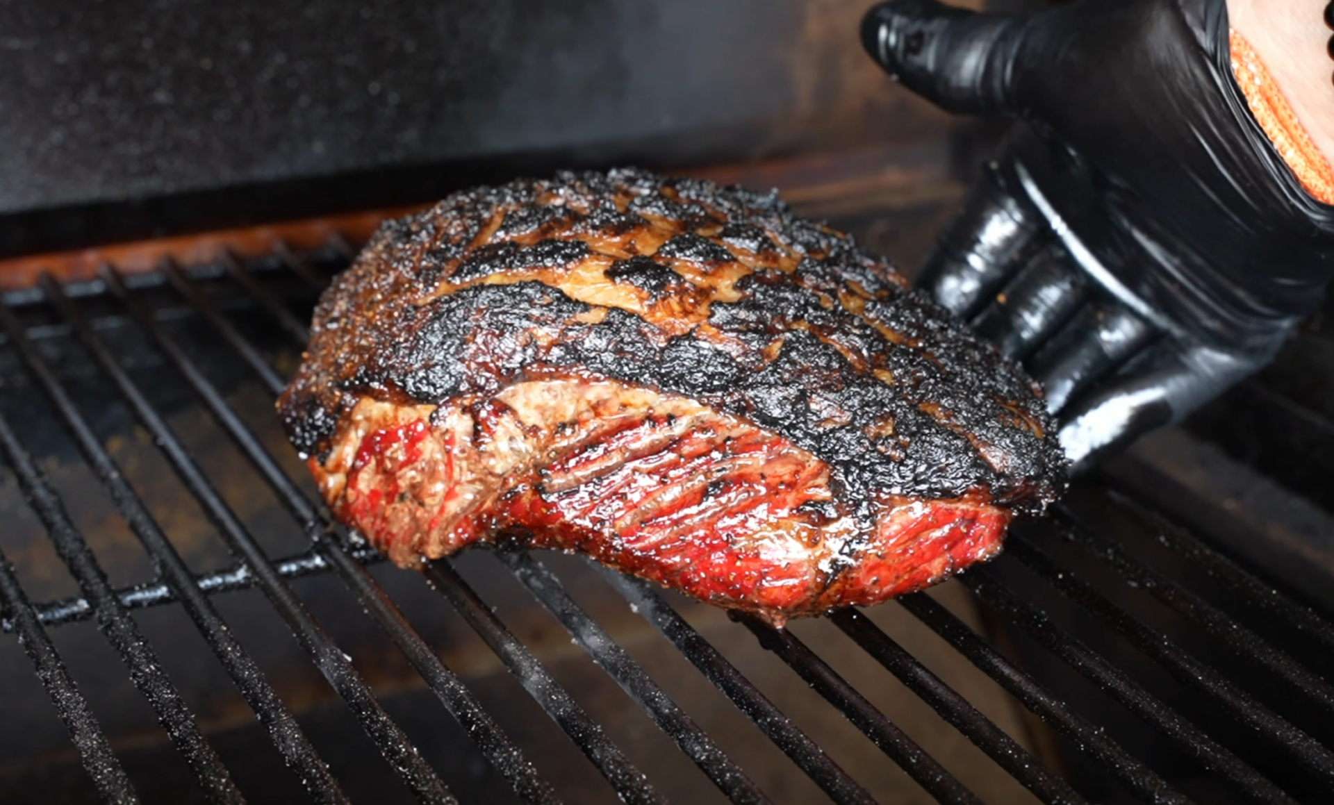 A gloved hand checks a large, charred brisket with a caramelized crust on a metal grill inside a smoker, with grill marks visible and a smoky background.