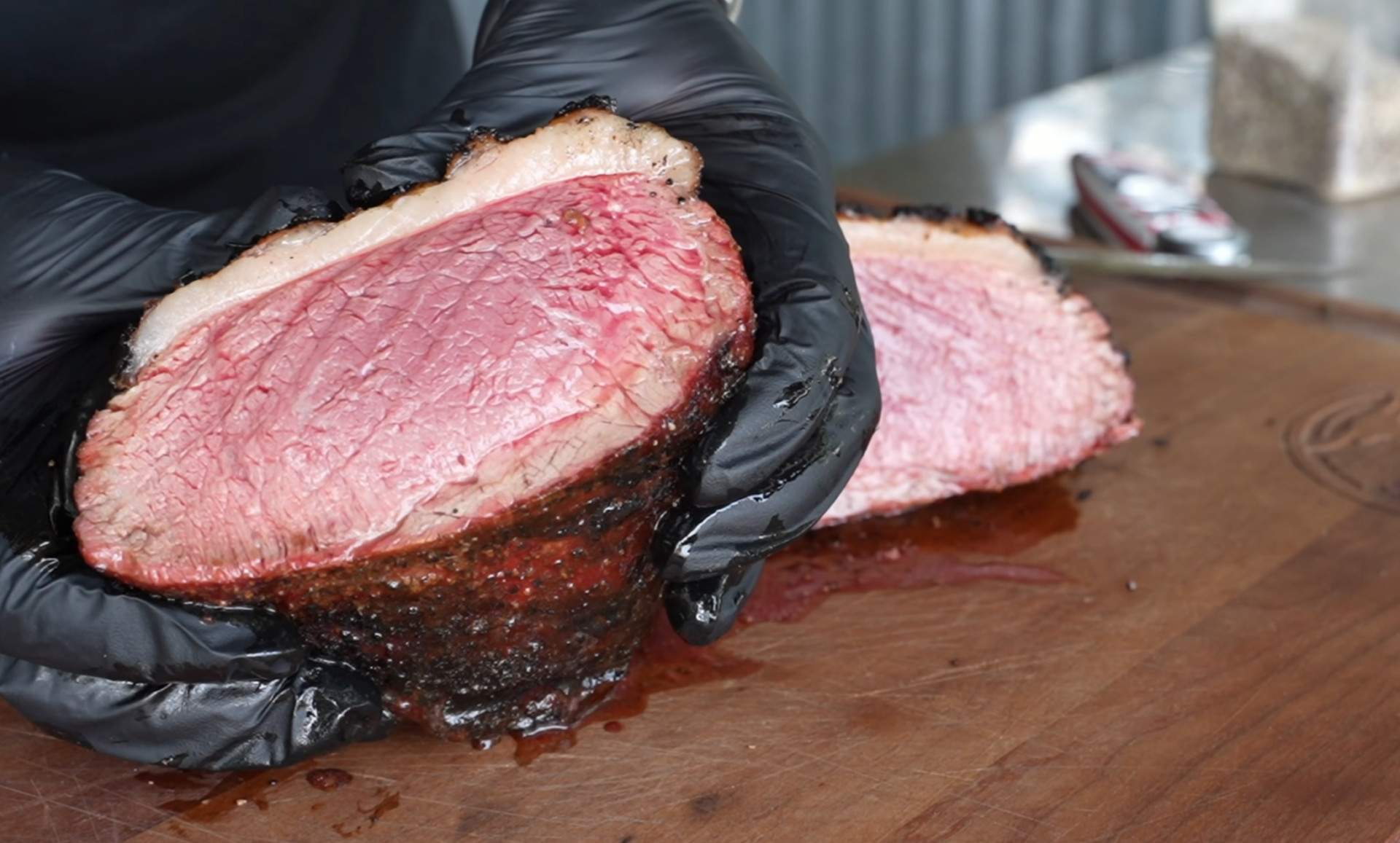 A person wearing black gloves holds a sliced, juicy, medium-rare roast beef with a charred crust on a wooden cutting board.