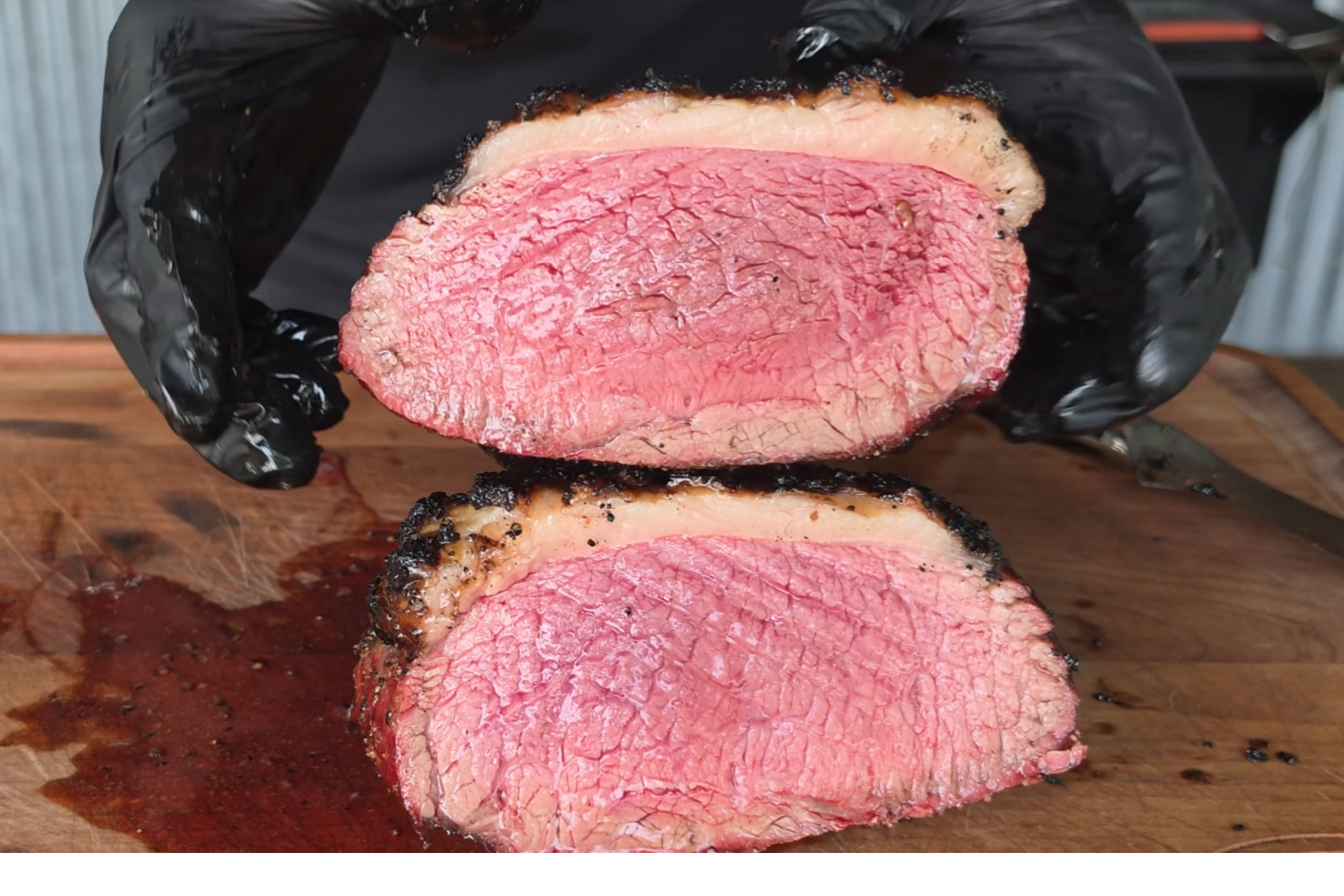 A person wearing black gloves holds two halves of a sliced, juicy piece of rare cooked beef with a seared crust, displayed on a wooden cutting board.