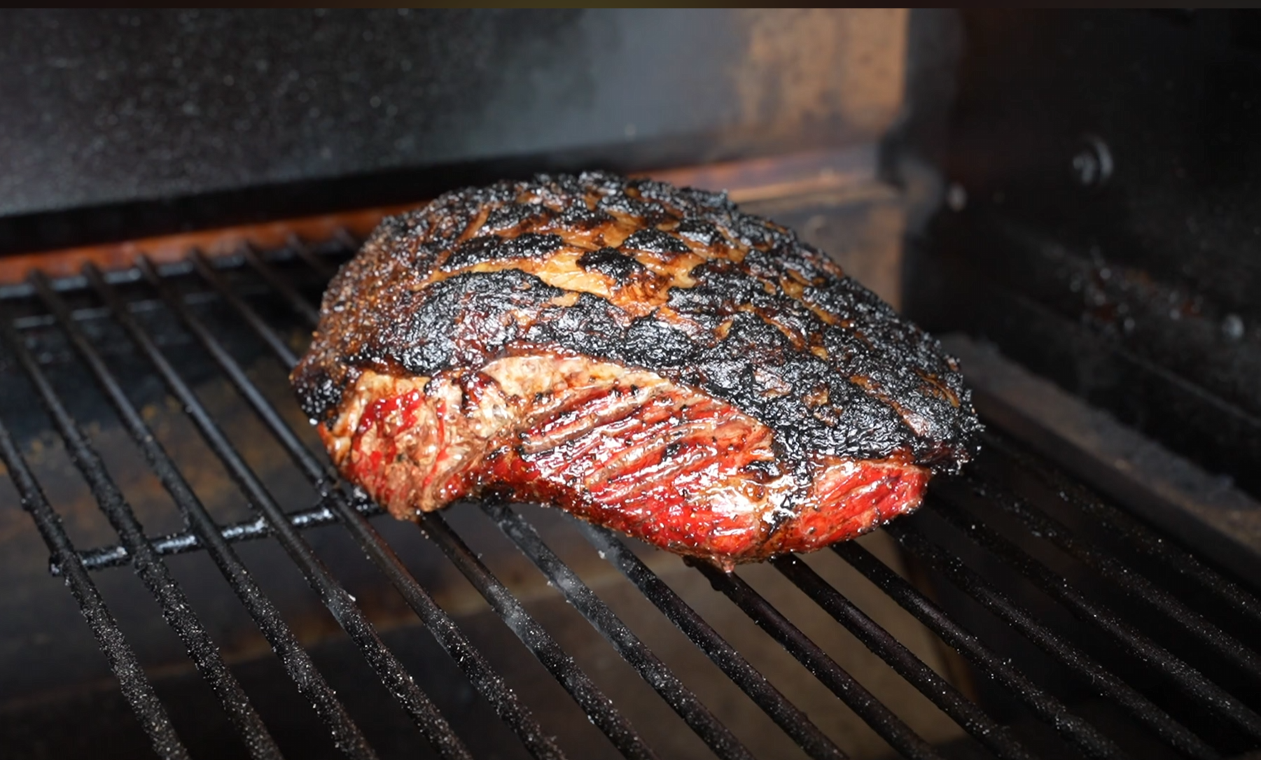A smoked brisket with a dark, crispy bark sits on a grill rack inside a smoker. The meat appears juicy and tender, with visible grill marks and charred edges.