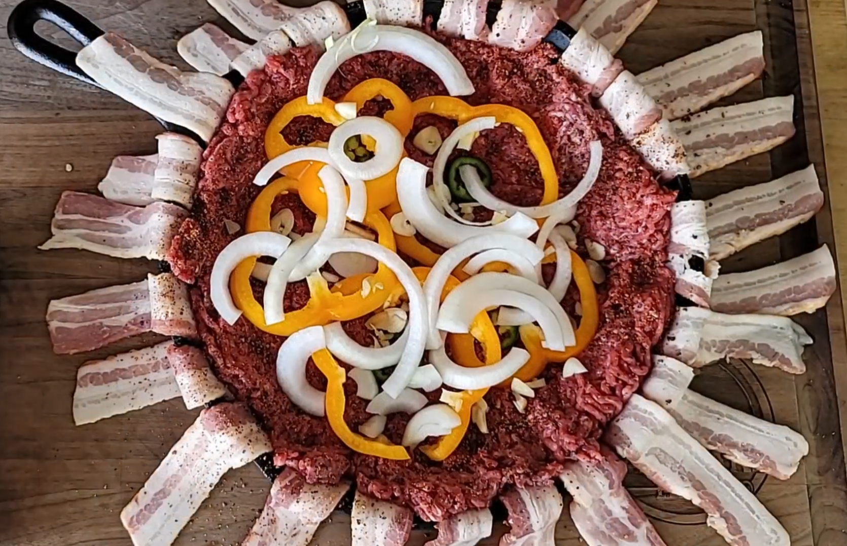 A cast iron pan lined with raw bacon strips, filled with ground beef, and topped with sliced yellow bell peppers and onions, sits on a wooden surface.