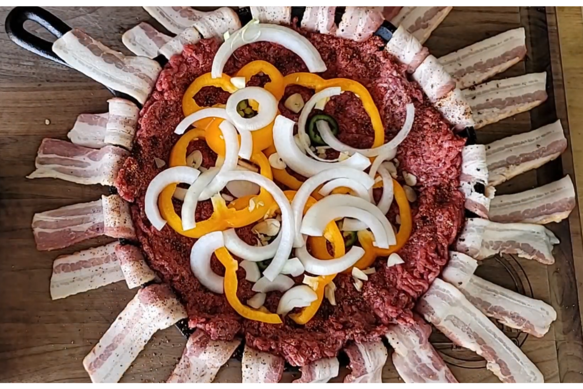 A round pan with raw ground beef, lined with bacon strips, topped with sliced yellow bell peppers, onions, and garlic, ready to be cooked.
