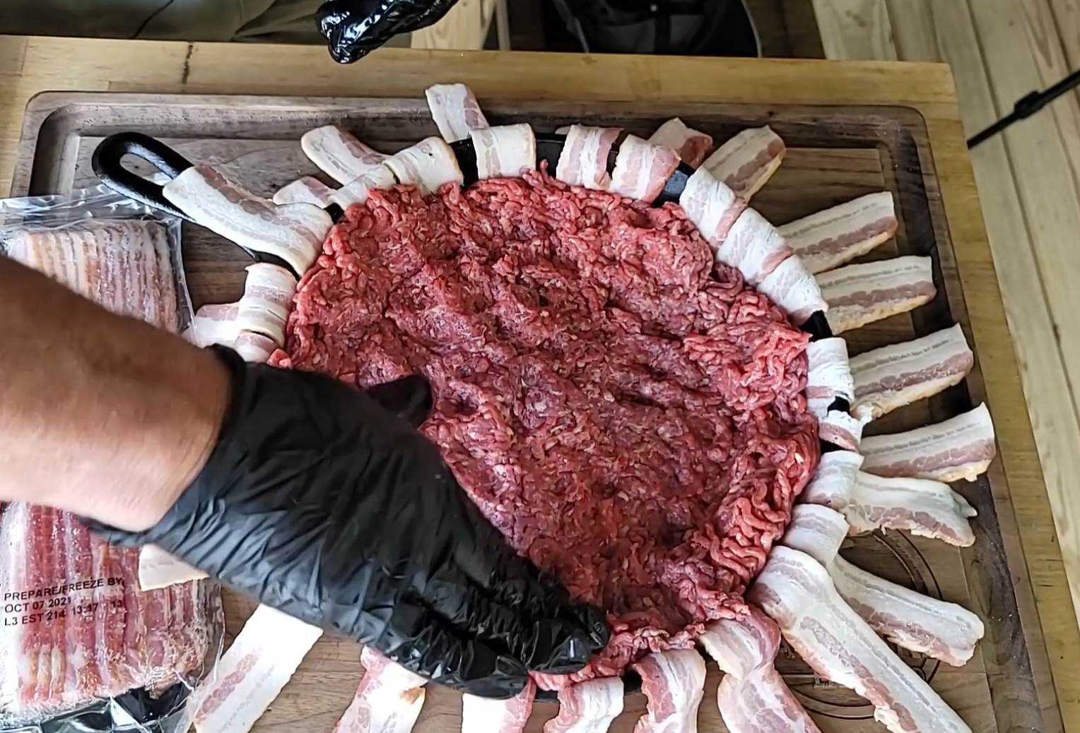 A person wearing black gloves spreads ground beef over a wooden cutting board lined with strips of bacon, preparing a meat dish. A packaged bacon strip is visible on the side.