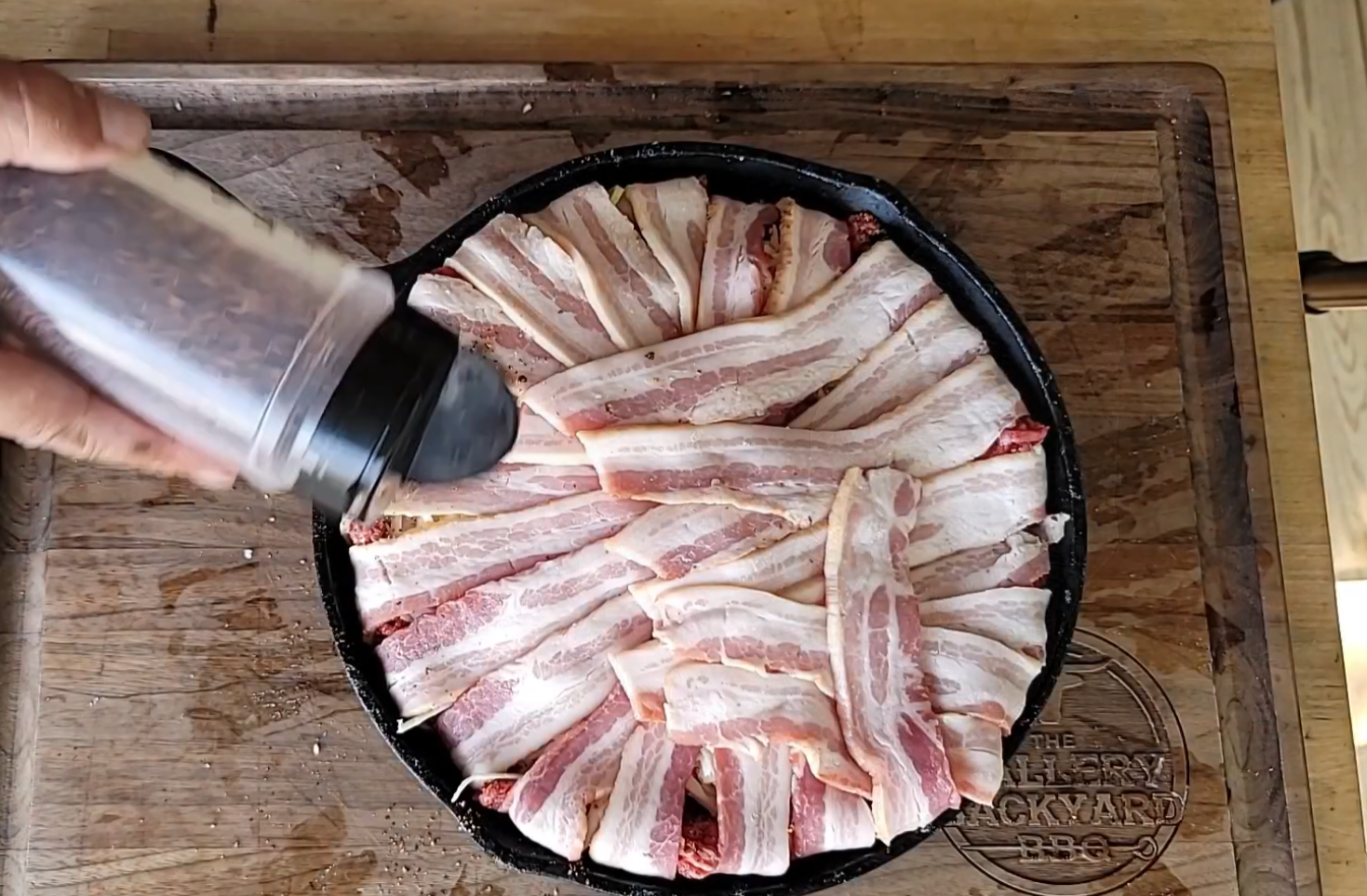 A hand sprinkles seasoning over a cast iron pan filled with strips of raw bacon arranged in a crisscross pattern on a wooden cutting board.