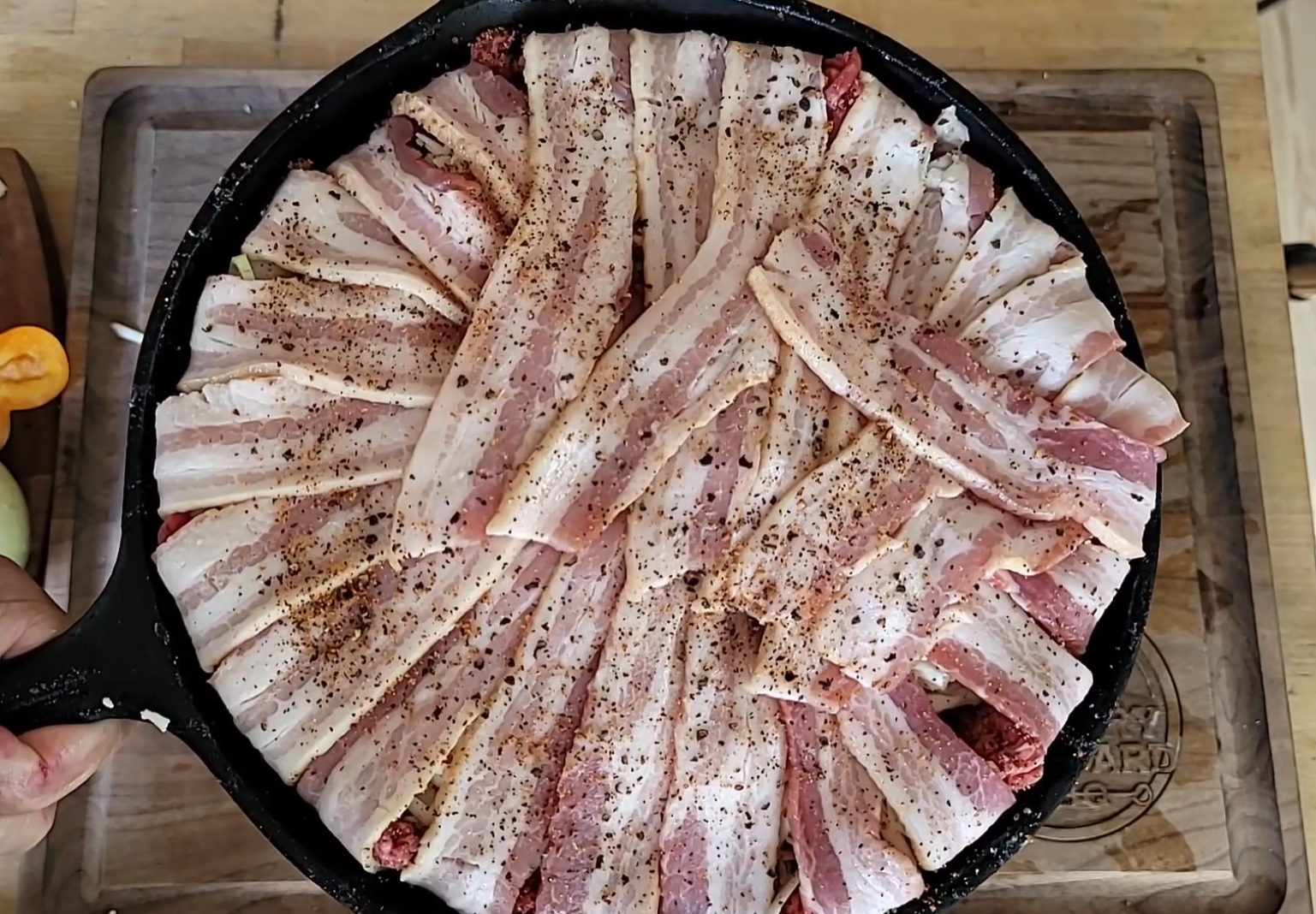 A cast iron skillet filled with raw bacon strips arranged in a circular pattern over a dish, sprinkled with black pepper, is held above a wooden cutting board.