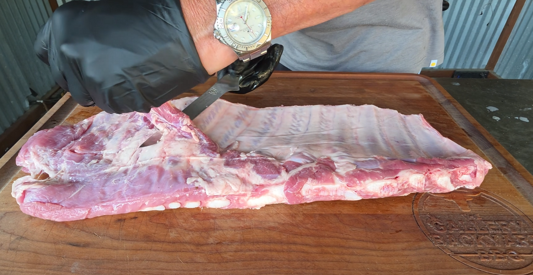 A person wearing a black glove uses a knife to trim the membrane from a rack of raw pork ribs on a wooden cutting board.