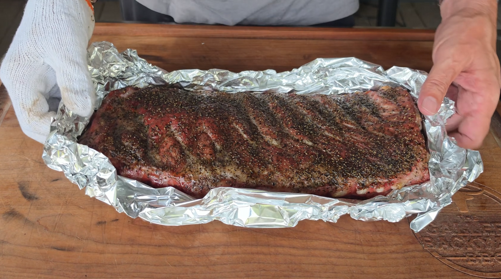 A person wraps a seasoned rack of ribs in aluminum foil on a wooden surface, preparing the meat for cooking.