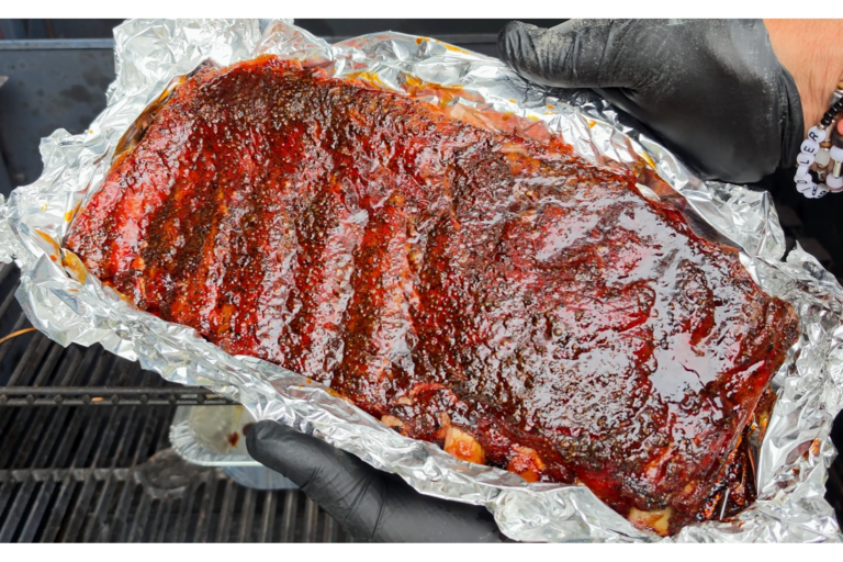 A rack of glazed, barbecued ribs sits on crinkled foil, held by gloved hands above a grill. The ribs appear juicy and caramelized with a rich, dark red barbecue sauce.