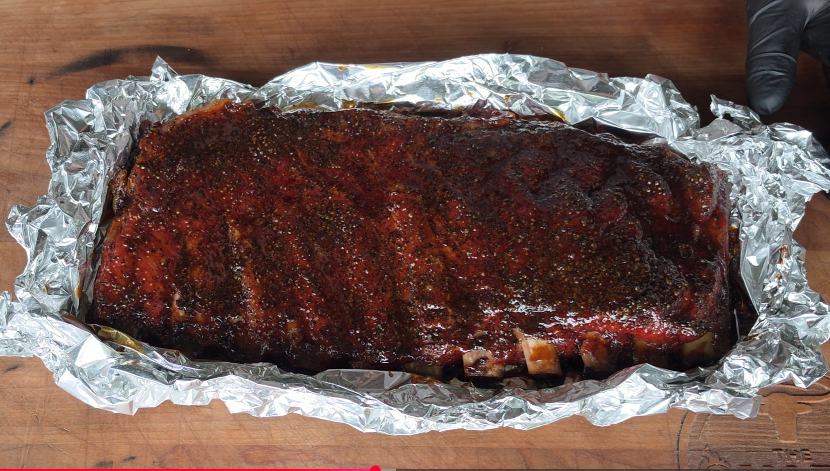 A rack of seasoned, glazed ribs sits on a sheet of aluminum foil atop a wooden surface, with a gloved hand visible in the corner.