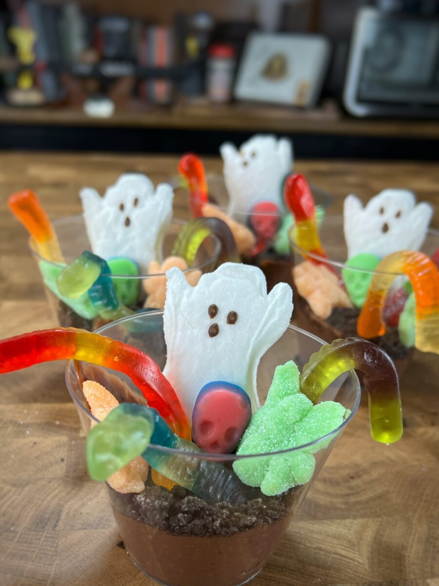 Plastic cups filled with chocolate pudding, topped with cookie crumbs, gummy worms, colorful candies, and marshmallow ghost-shaped treats arranged for a festive Halloween dessert.