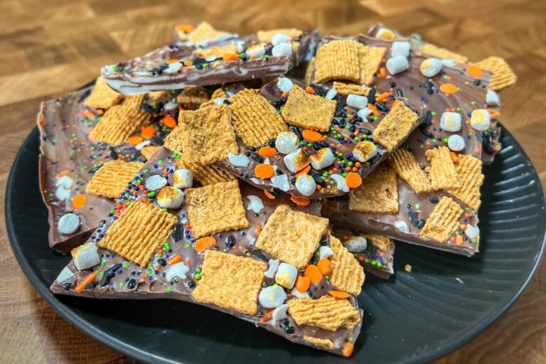 A plate of chocolate bark topped with colorful sprinkles, cereal squares, and mini marshmallows sits on a wooden surface.