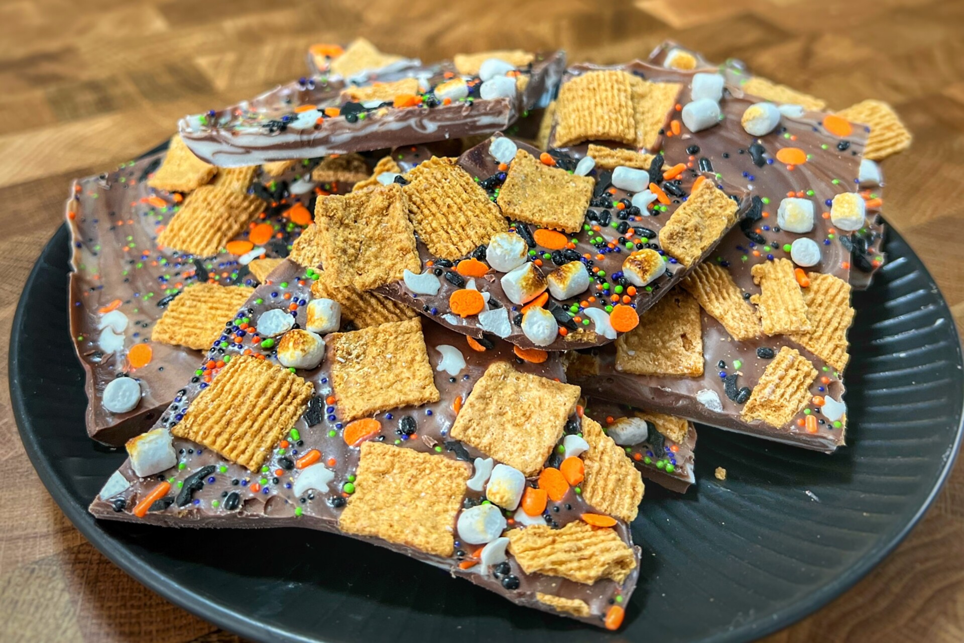 A plate of chocolate bark topped with colorful sprinkles, cereal squares, and mini marshmallows sits on a wooden surface.