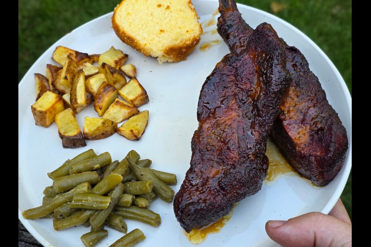 A plate with two pieces of barbecued meat, roasted potato cubes, green beans, and a slice of cornbread. The plate is held outside with grass in the background.