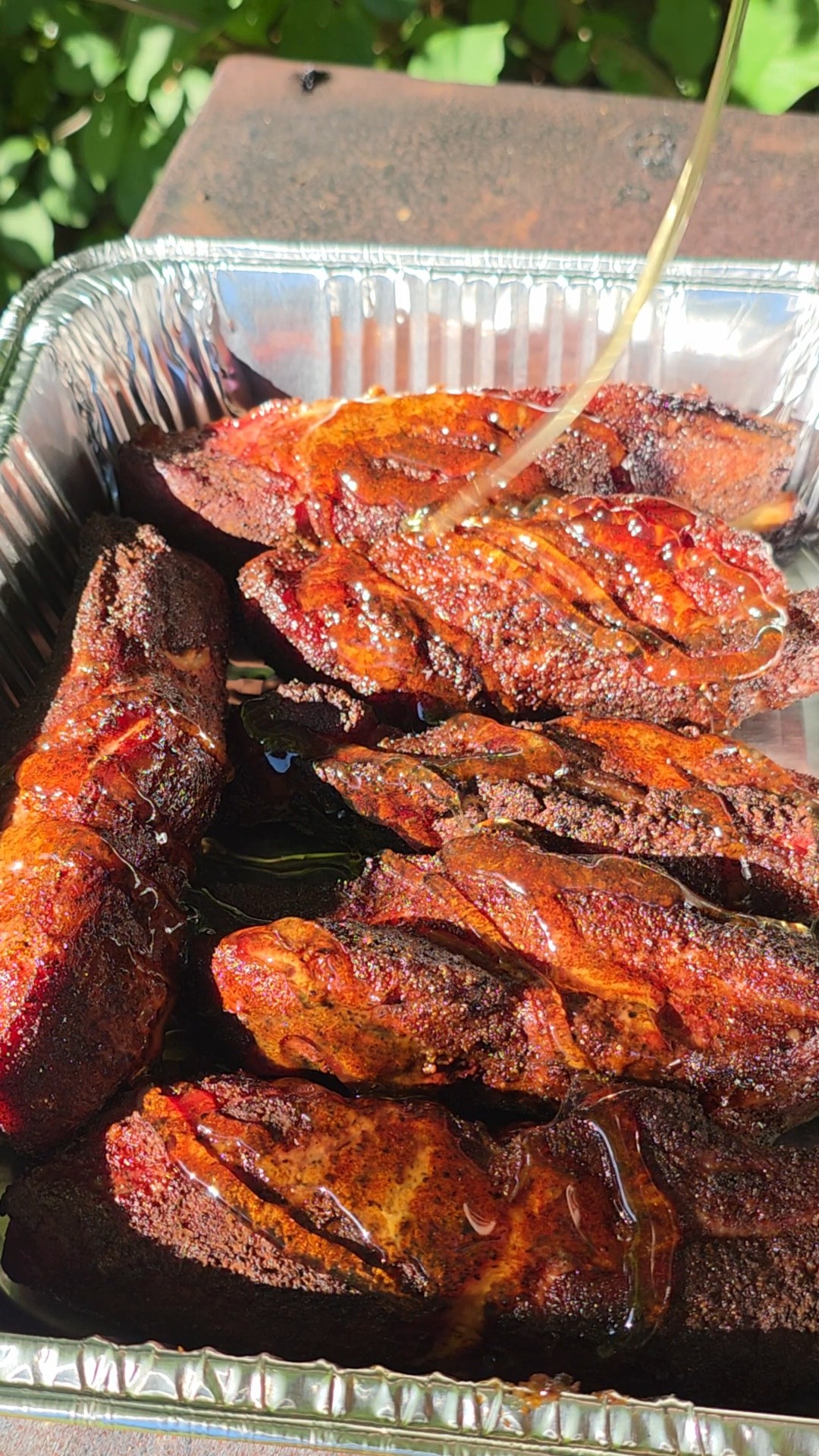 A close-up of barbecued ribs in a foil pan, being drizzled with a liquid glaze, with sunlight highlighting their crispy, seasoned surface. Green leaves are visible in the blurred background.