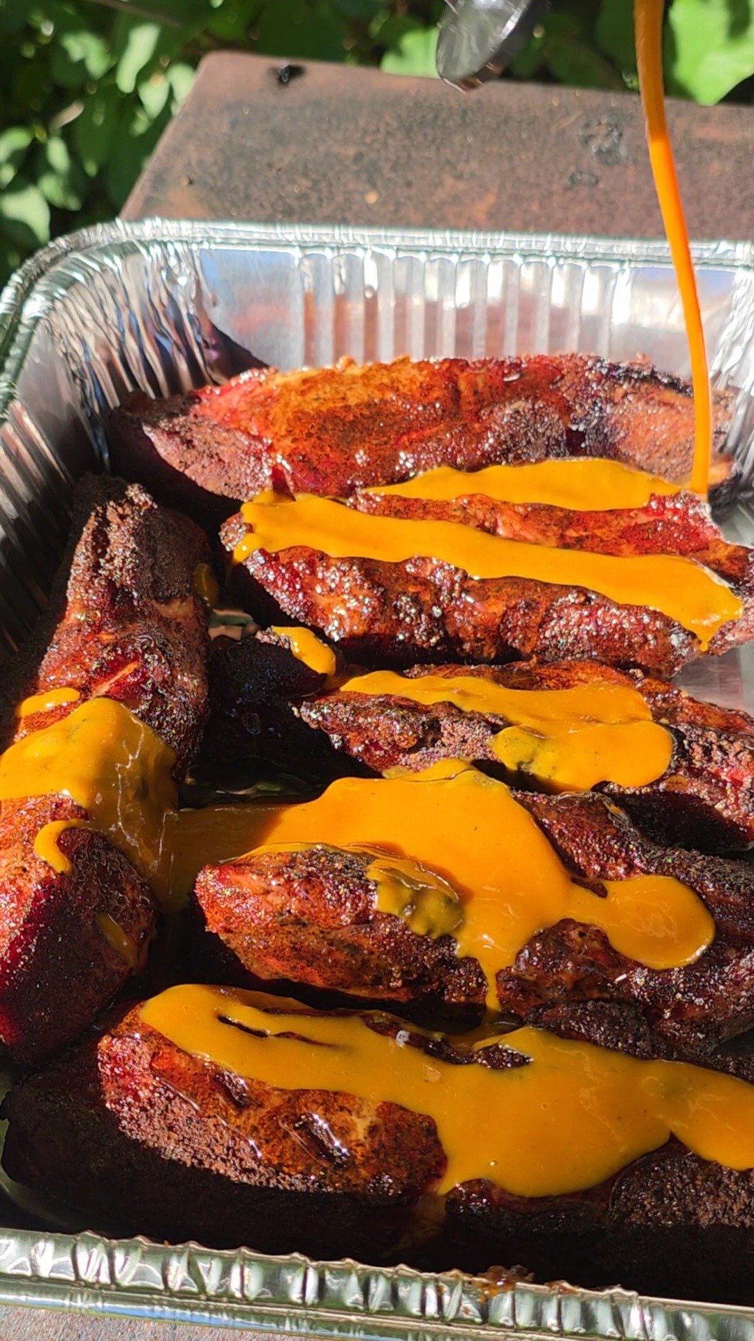 Barbecue ribs in a foil tray are topped with a generous drizzle of orange sauce, with sunlight highlighting the glossy finish. Green leaves are visible in the blurry background.
