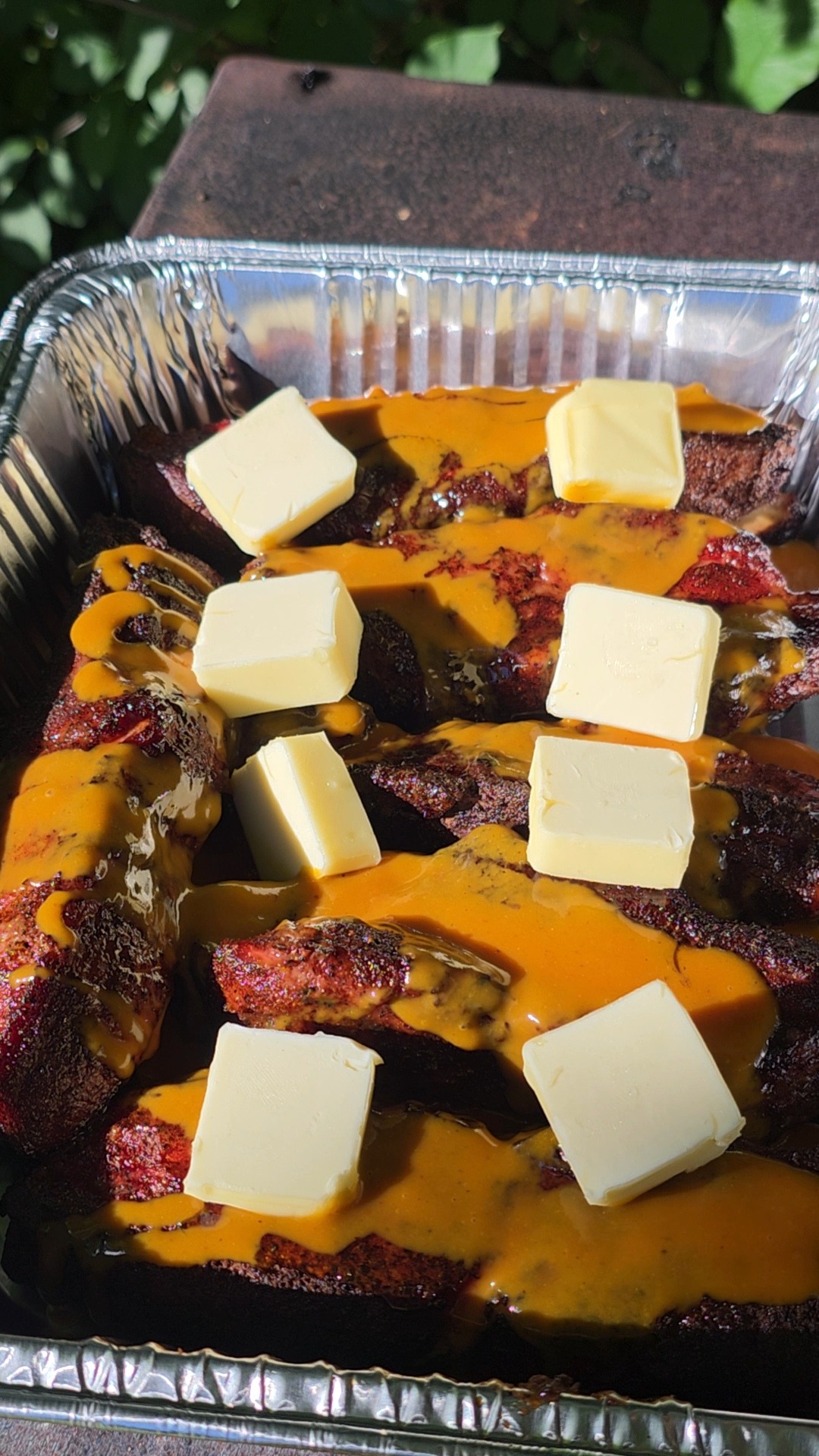 A foil tray filled with ribs covered in orange sauce, topped with several square pats of butter, outdoors with green leaves in the background.