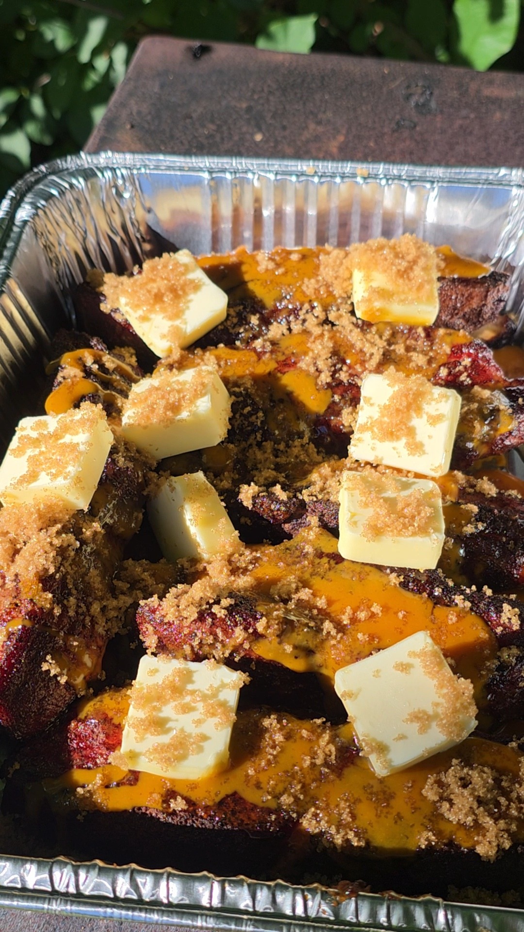 A foil tray filled with cooked plantains topped with melted cheese, cubes of butter, and sprinkled brown sugar, shown outdoors in sunlight.