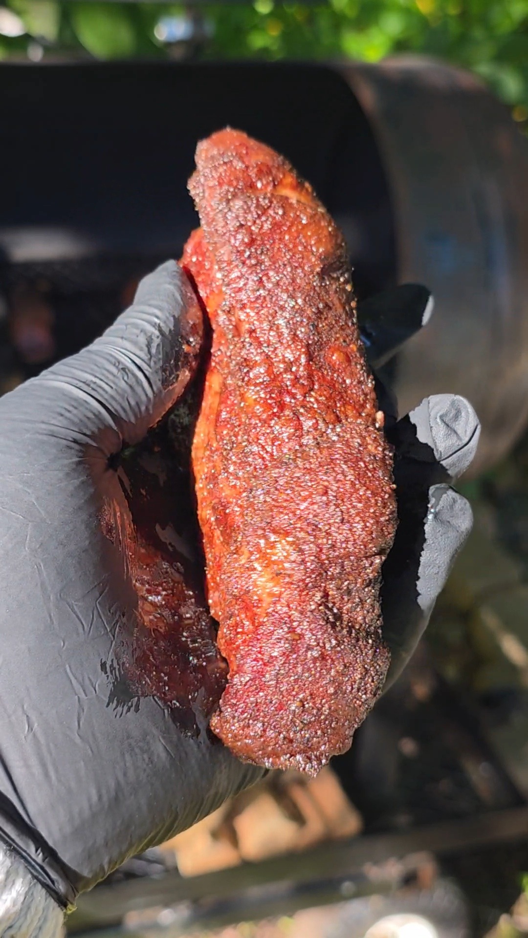 A gloved hand holds a seasoned, cooked piece of meat in front of a barbecue smoker outdoors, highlighting its crispy, textured surface.