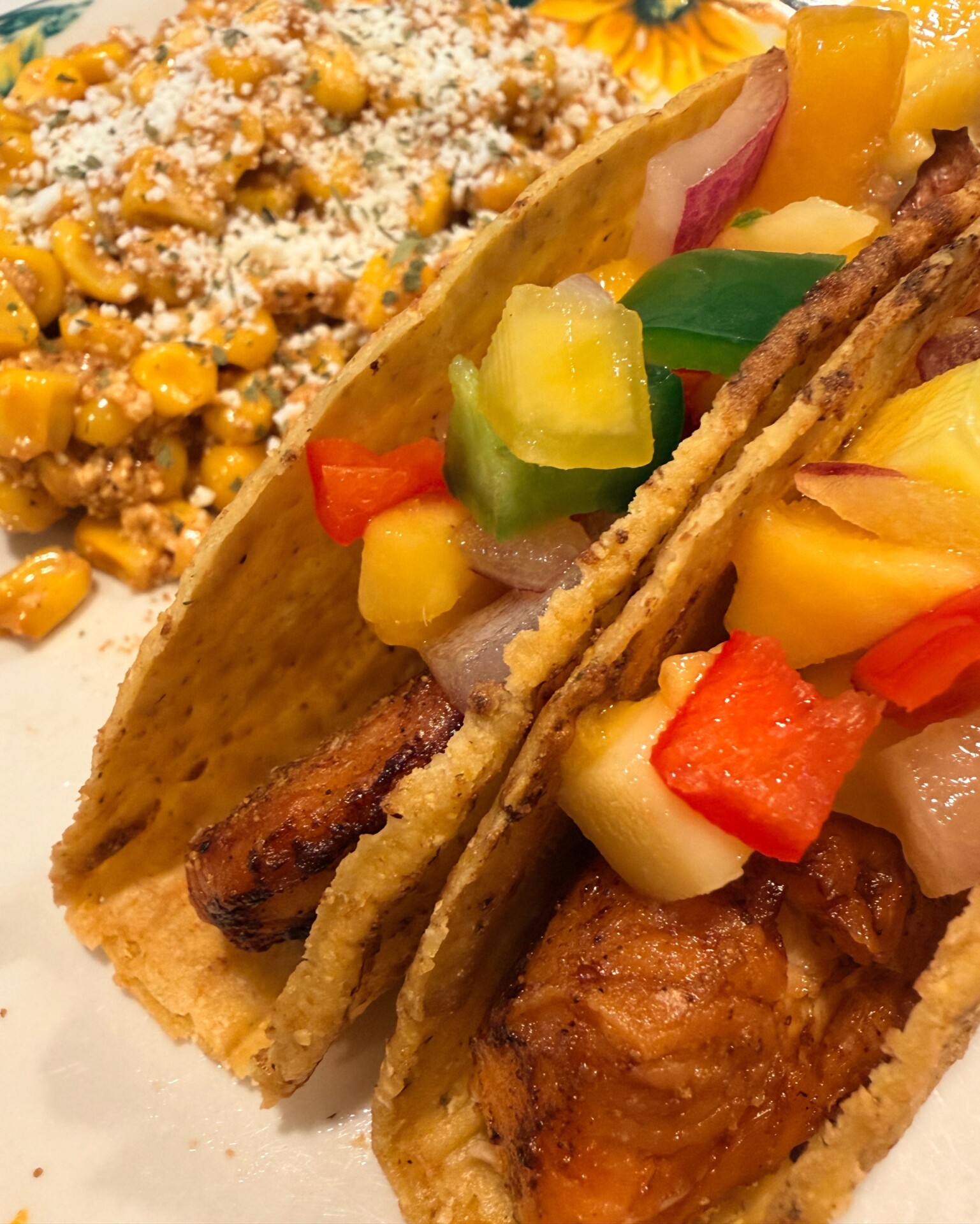 Two tacos with grilled fish and colorful pineapple salsa in crunchy shells, served with a side of Mexican street corn topped with cheese.
