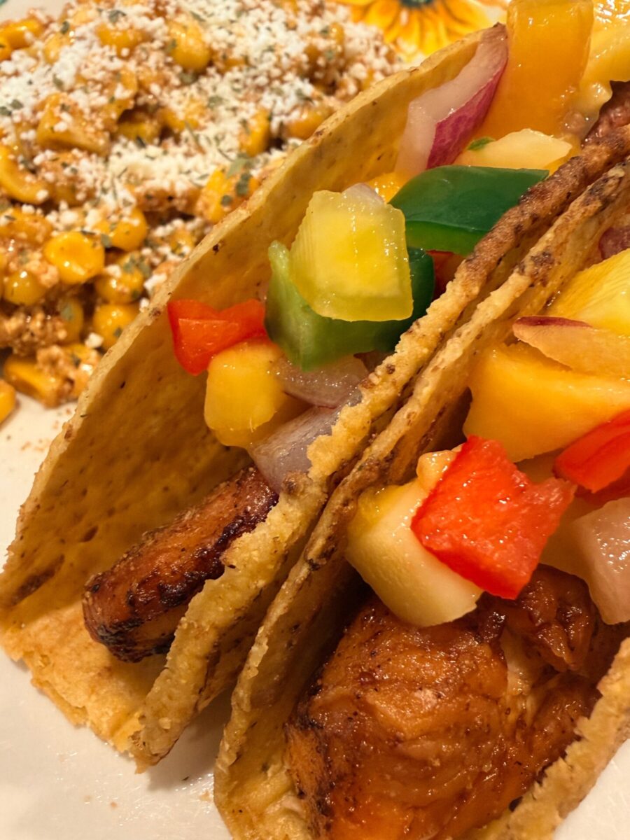 Two hard shell tacos filled with grilled salmon, diced pineapple, and colorful bell peppers, served next to a side of Mexican street corn topped with cheese and spices.