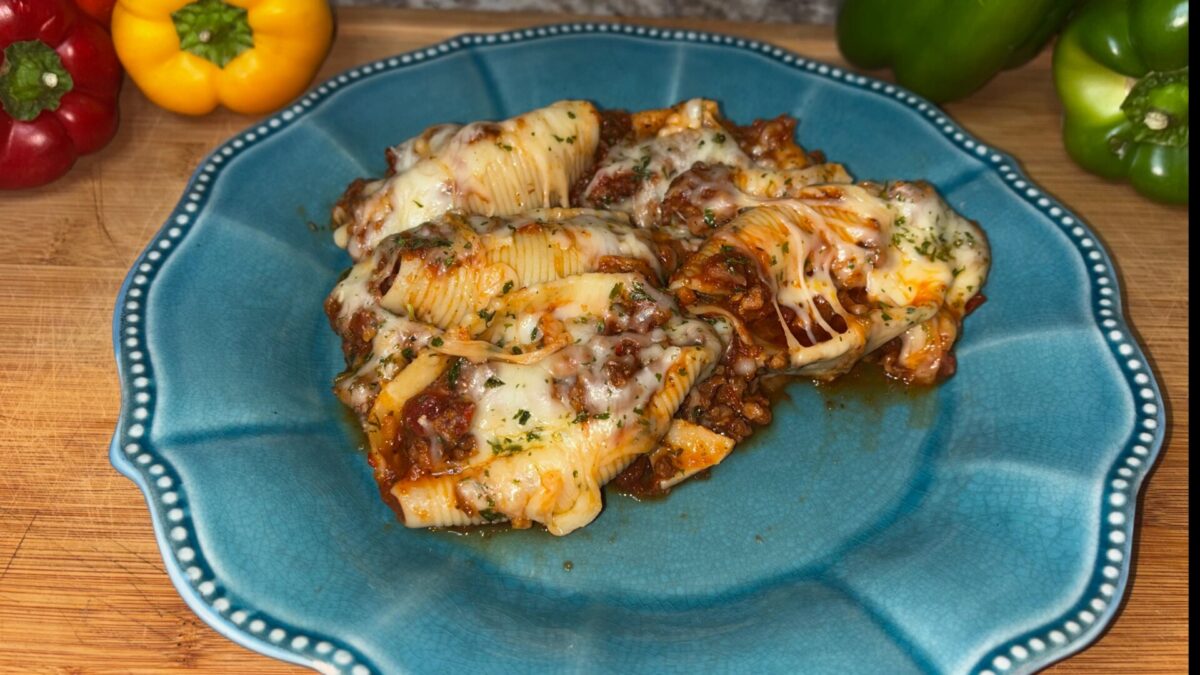 Stuffed pasta shells filled with meat and topped with melted cheese and herbs, served on a blue plate. Yellow, red, and green bell peppers are in the background.