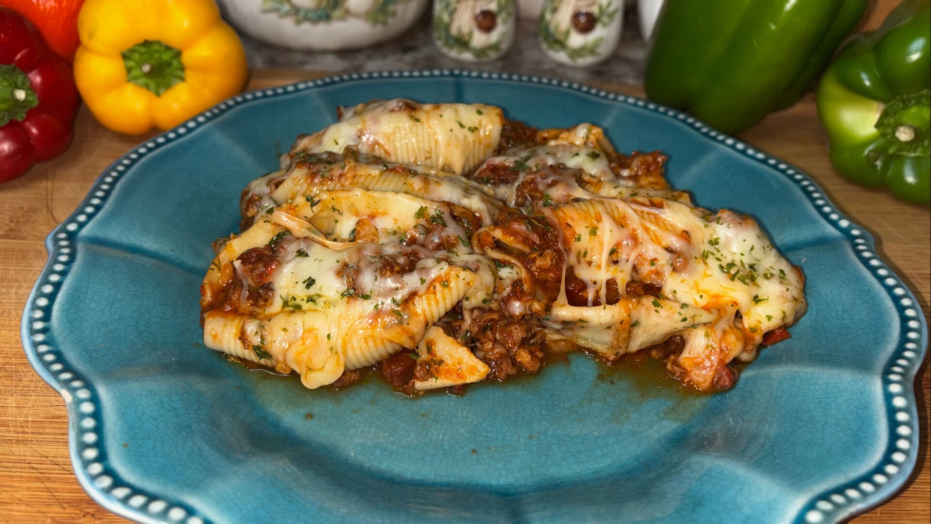 Stuffed pasta shells topped with melted cheese and herbs are served on a blue plate. Bell peppers in red, yellow, and green are seen in the background on a wooden surface.