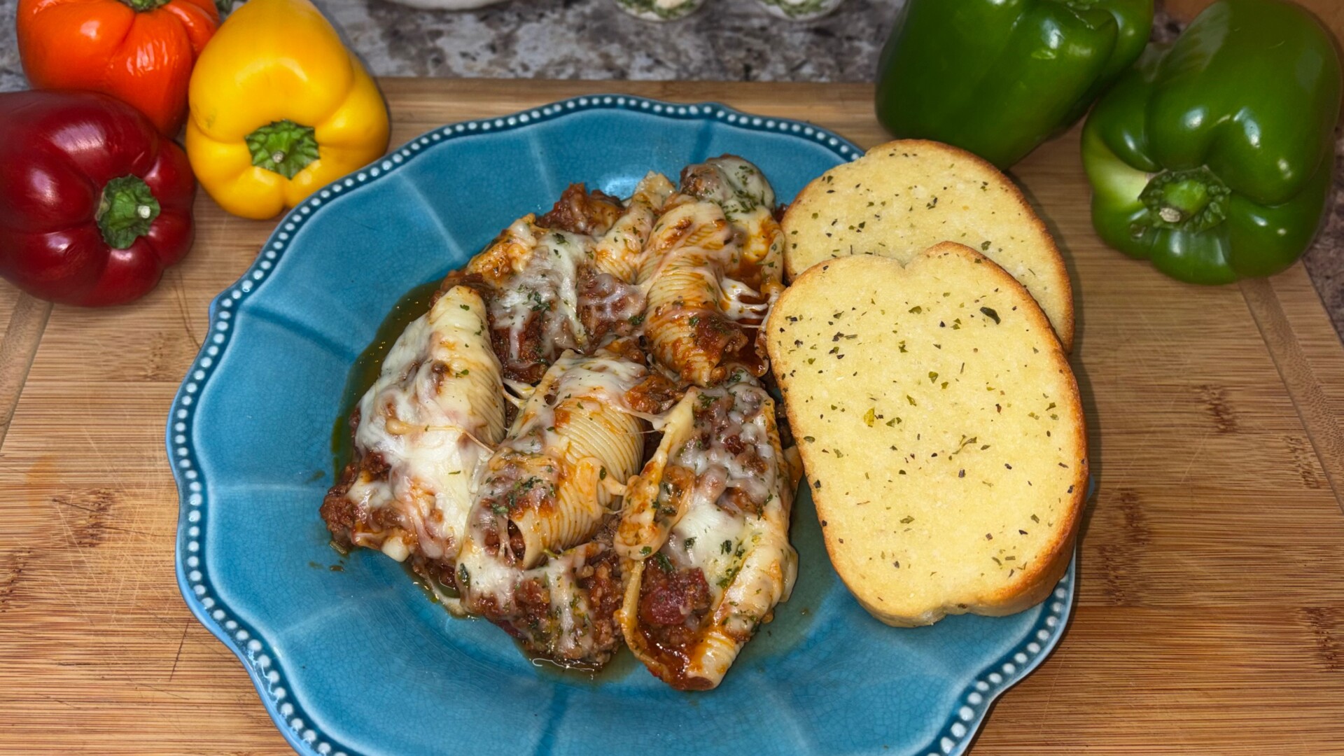 A blue plate with cheesy stuffed pasta shells and two slices of garlic bread, placed on a wooden board, surrounded by red, yellow, and green bell peppers.