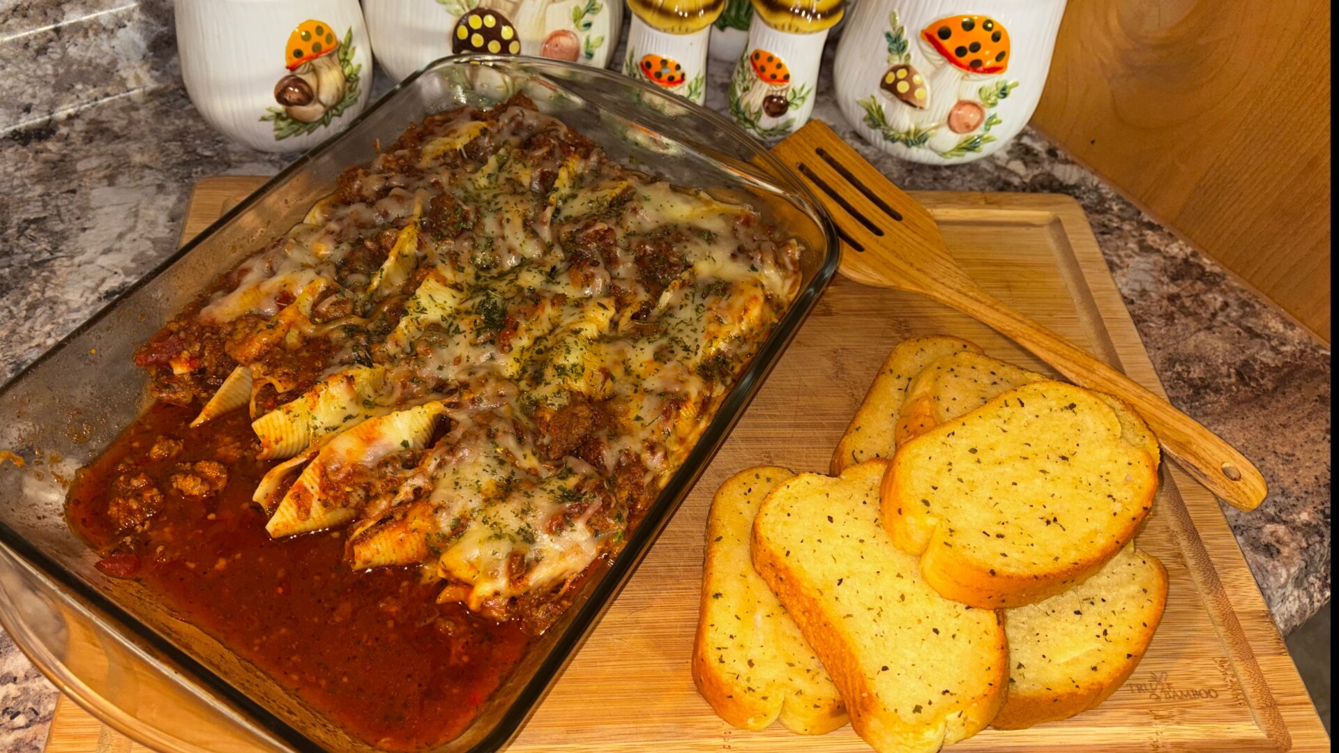A glass dish of baked pasta shells with meat sauce and melted cheese sits on a wooden board next to slices of garlic bread, with decorative jars in the background.