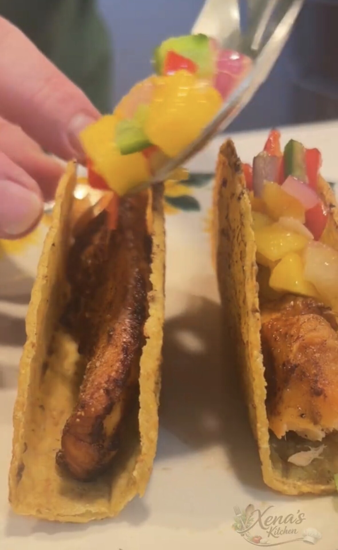 A hand tops two fish tacos in corn tortillas with colorful mango salsa using a spoon. The tacos are placed side by side on a white plate, and Xenas Kitchen is subtly visible in the lower right corner.