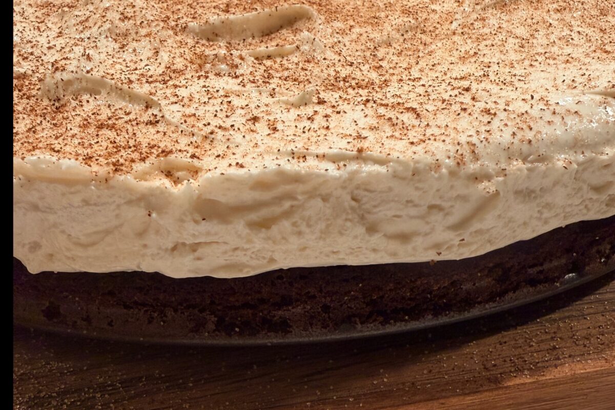 A close-up of a layered dessert with a thick, creamy white topping dusted with cocoa powder, sitting on a dark chocolate base on a wooden surface.