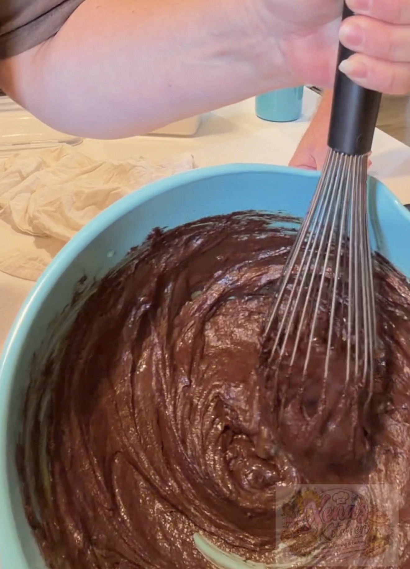 A person whisking thick chocolate batter in a large blue bowl, with a napkin and a cup visible in the background.