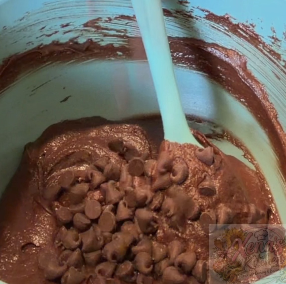 A light blue spatula stirs chocolate chip cookie dough in a matching bowl. A cluster of chocolate chips is visible on the thick, brown batter.