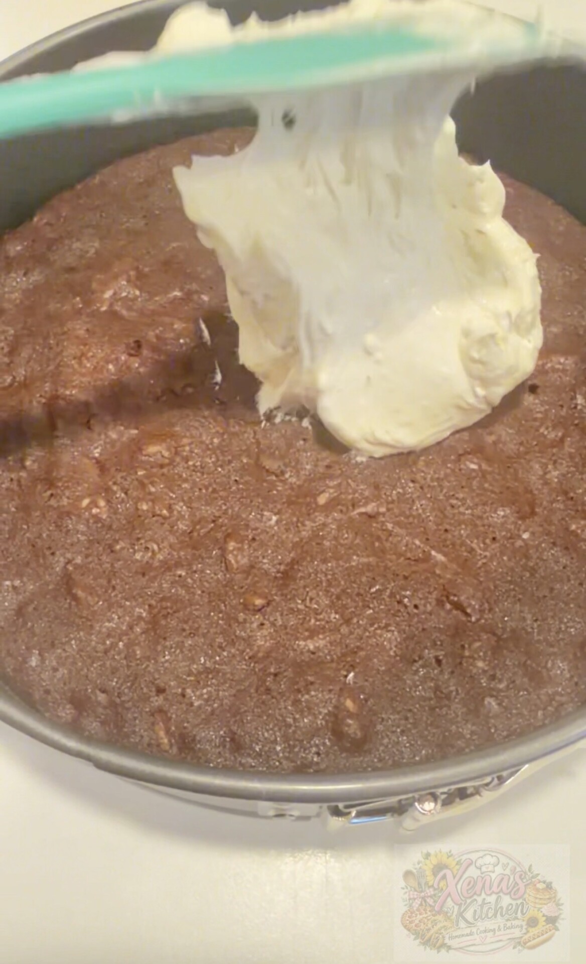 A layer of white frosting is being spread onto a round, baked chocolate cake in a metal pan. The image includes a logo reading Xenas Kitchen in the bottom right corner.