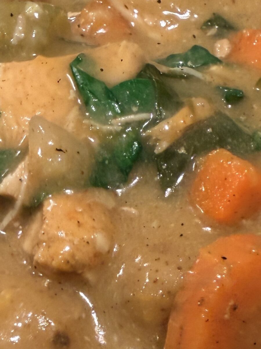 Close-up of a thick stew with chunks of chicken, carrots, potatoes, and leafy greens in a creamy, seasoned broth.