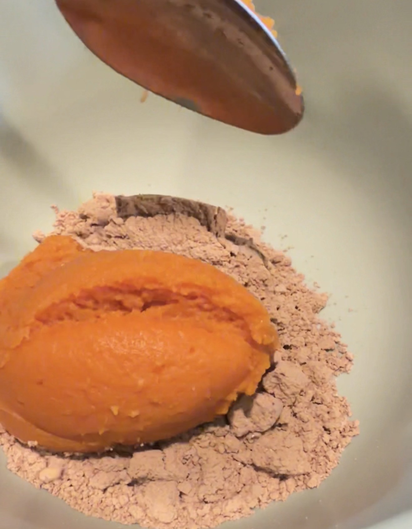 A metal spoon holds pumpkin puree above a bowl filled with cocoa powder, with a scoop of pumpkin puree already on top of the powder.