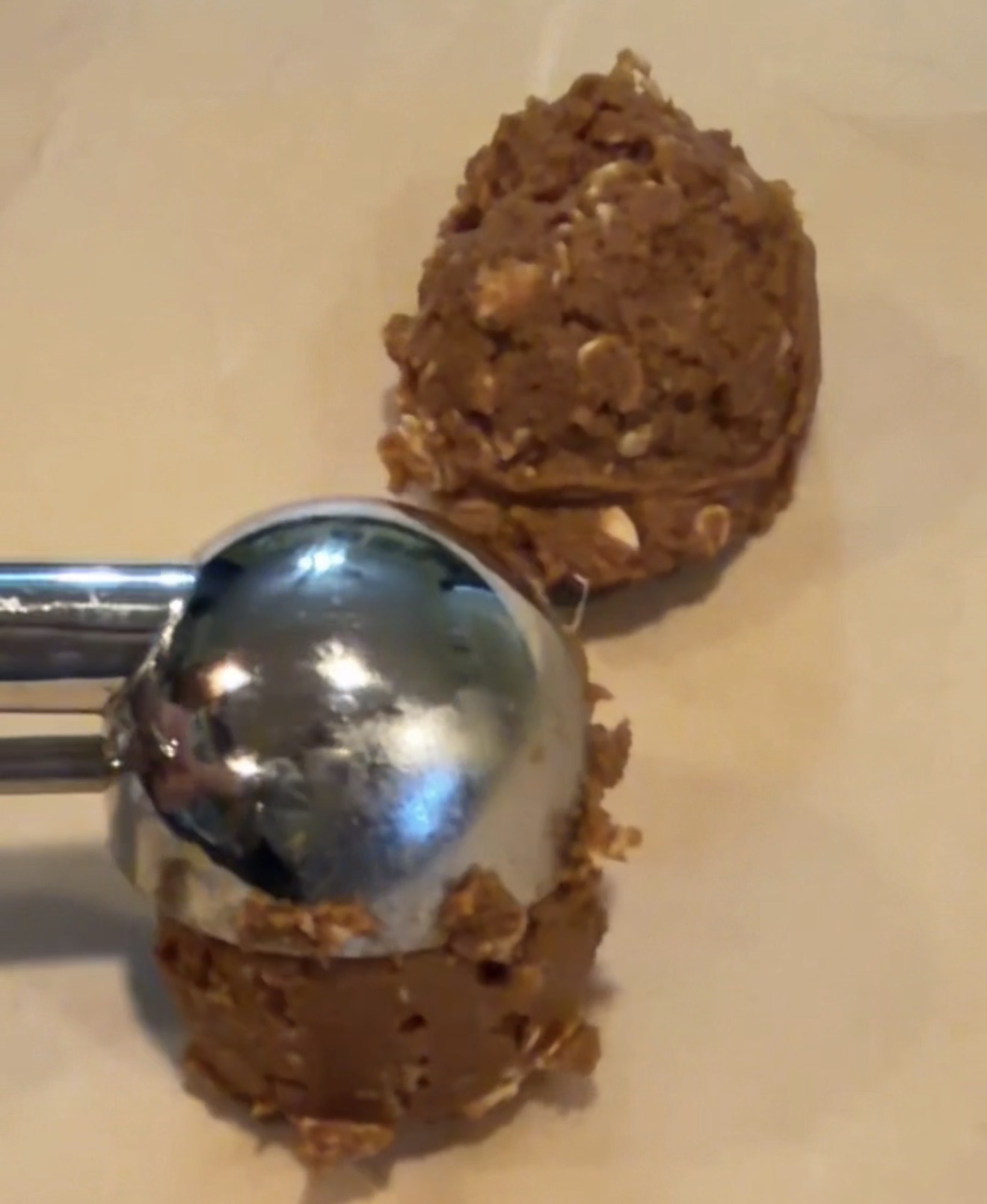 A metal cookie scoop holds a ball of chocolate oatmeal cookie dough, with another dough ball on parchment paper nearby, ready for baking.