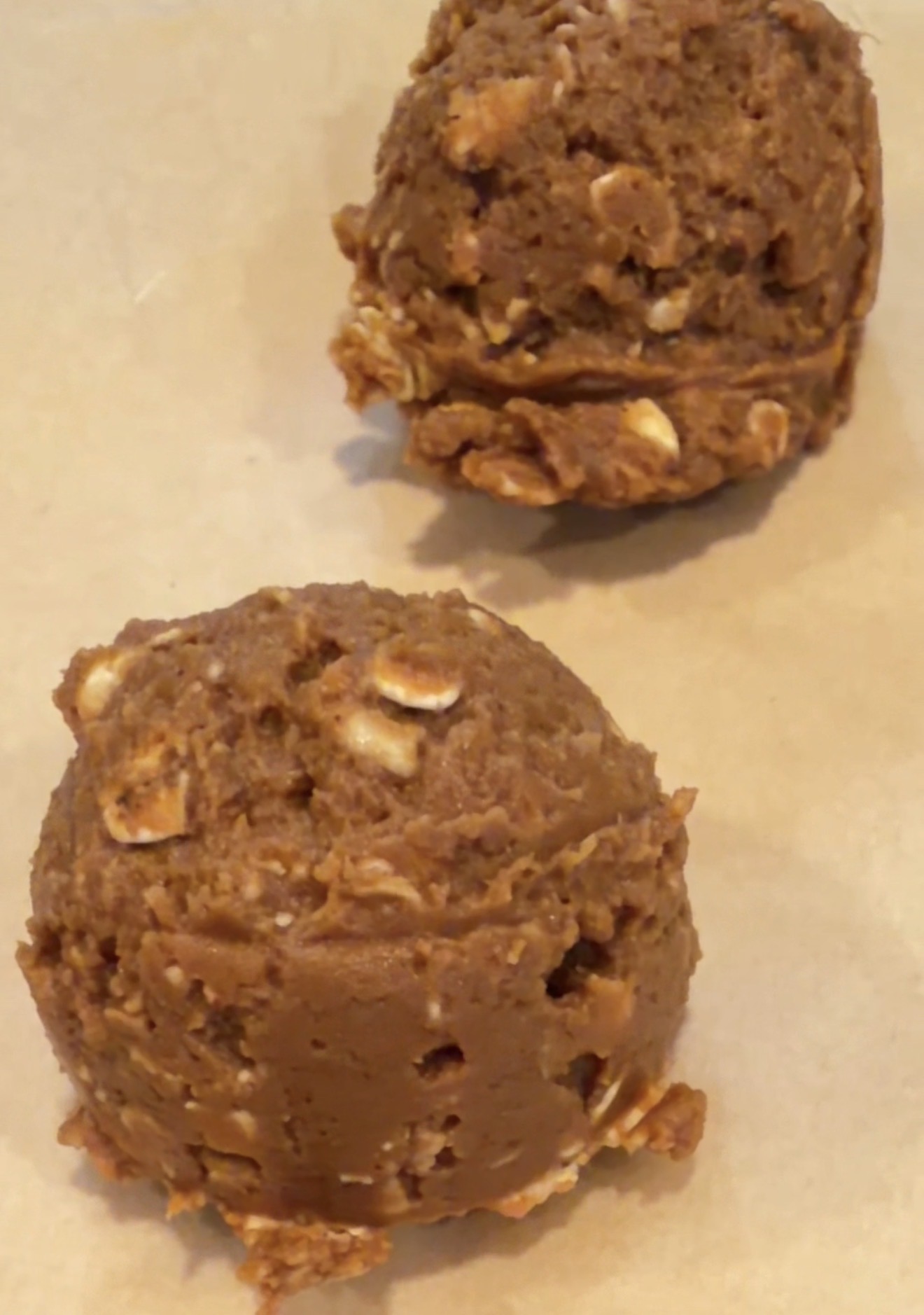 Two scoops of raw cookie dough with visible oats, placed on a sheet of parchment paper, ready to be baked.