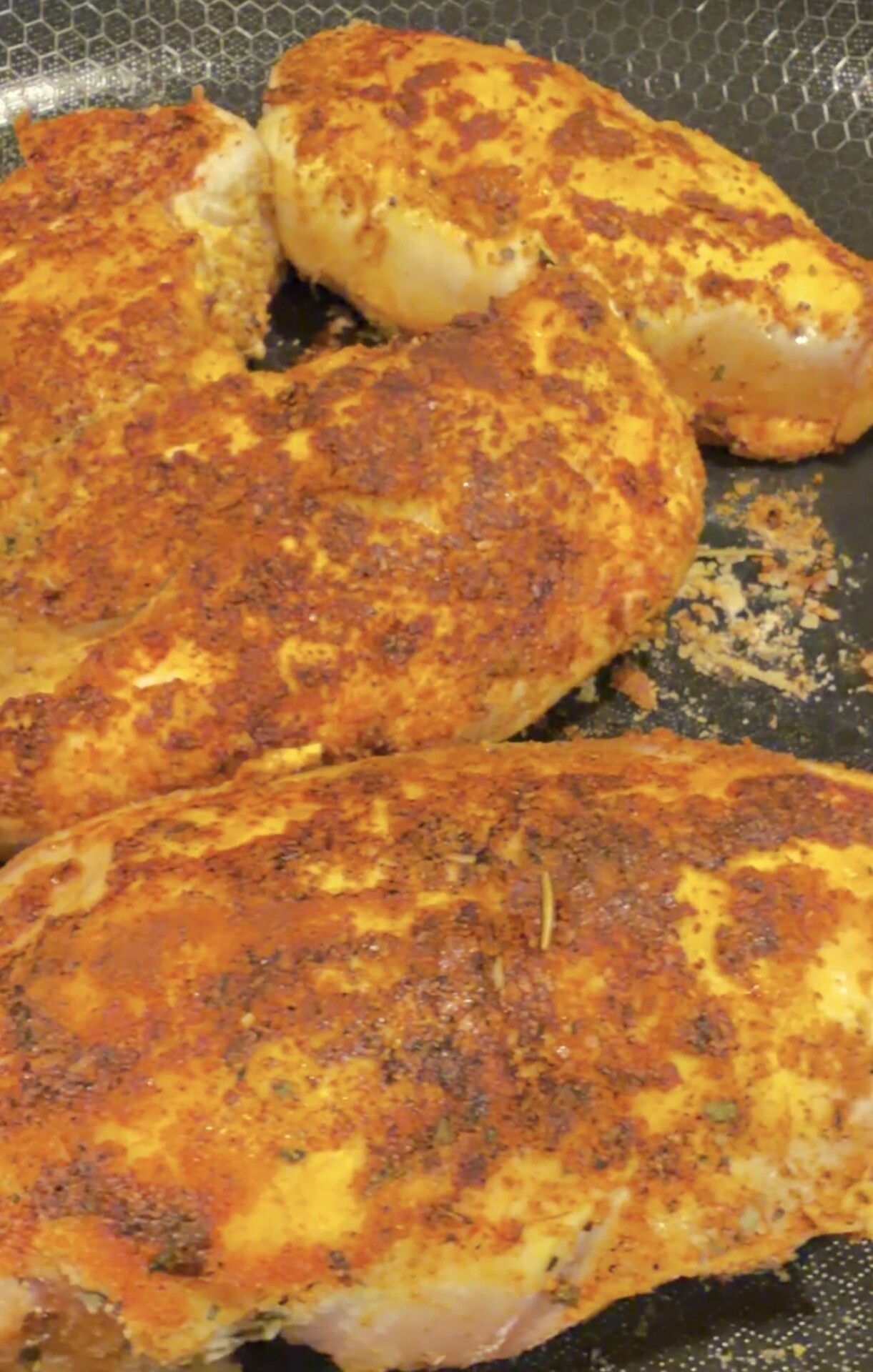 Seasoned chicken breasts cooking on a skillet, with a golden-brown, spiced crust forming on the surface. The background shows a textured, nonstick pan.