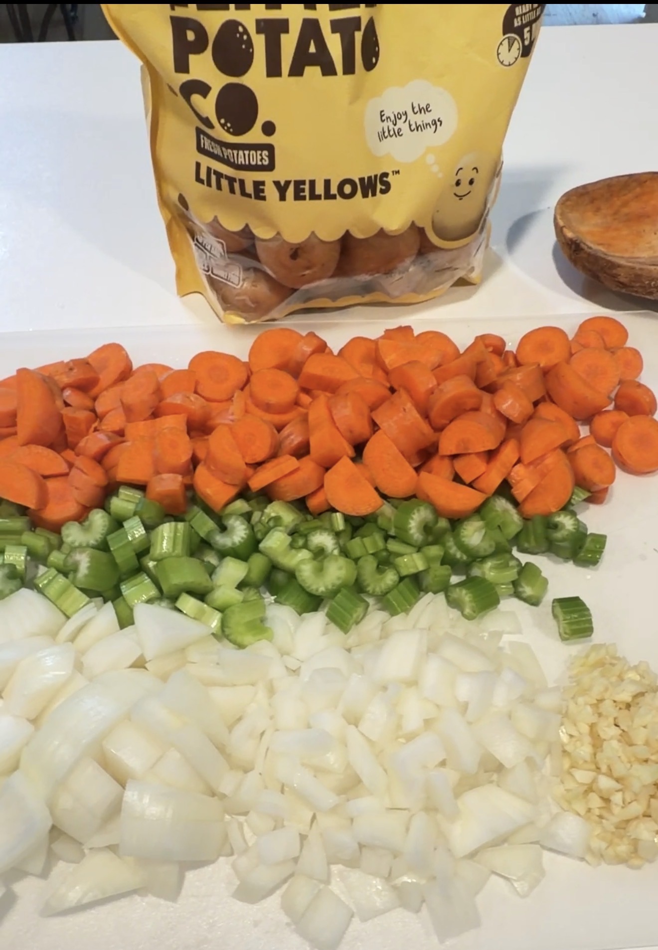 Chopped carrots, celery, onion, and garlic are arranged in rows on a cutting board. Behind them is a bag of Potato Co. Little Yellows potatoes on a white countertop.