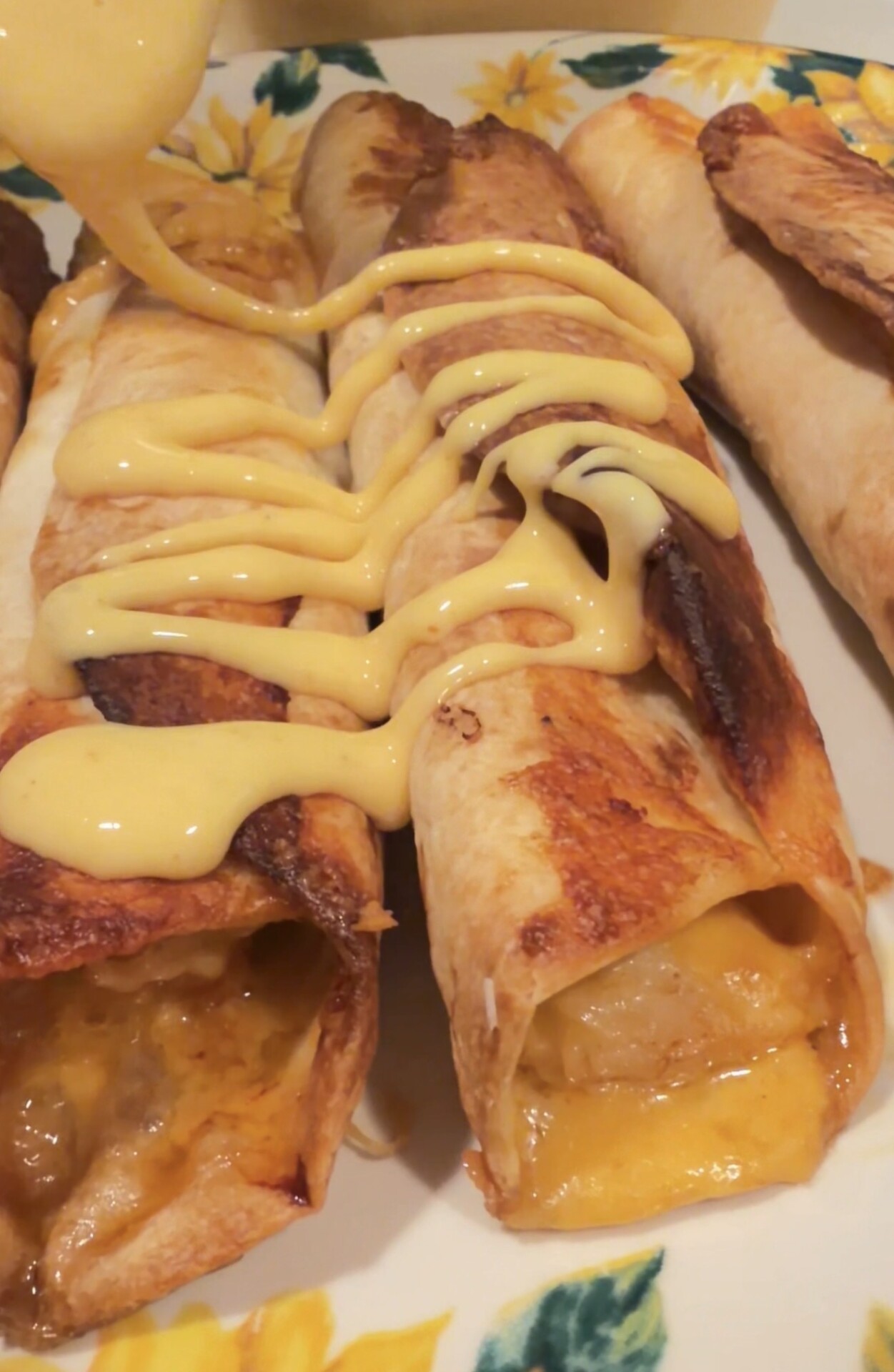 Three crispy rolled tortillas filled with melted cheese, served on a floral plate, being drizzled with creamy yellow cheese sauce.